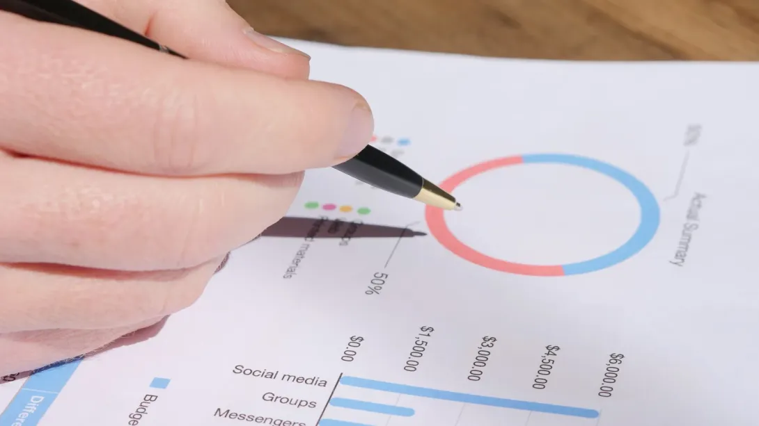 A hand with a pen reviewing a printed monthly budget report with charts